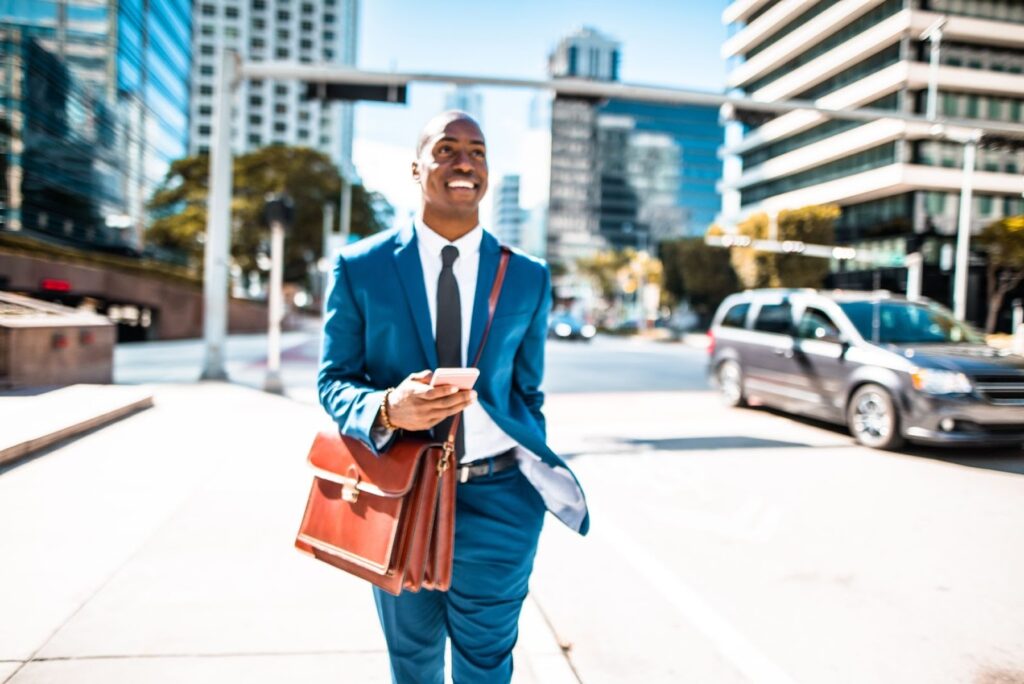 A man walks in a city setting while carrying a brown briefcase and holding a cell phone.